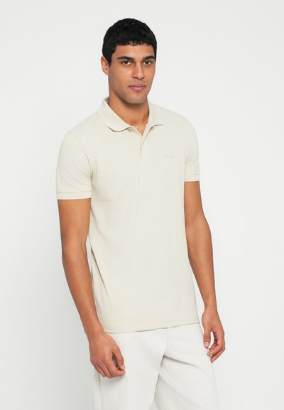 Polo beige avec une texture douce, manches courtes et patte de boutonnage à deux boutons. Présente un logo subtil en fil assorti sur la poitrine.