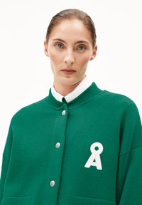 Cardigan tricoté vert avec un logo blanc, dotée de boutons-pression en argent, d'un col haut et de poches latérales, en tissu texturé.