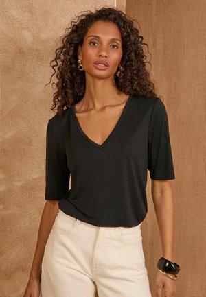 Frau mit lockigem Haar, die ein schwarzes V-Ausschnitt-Kurzarmoberteil, beigefarbene Hose, goldene Creolen und schwarze sowie goldene Armbänder vor braunem Hintergrund trägt.