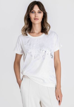 Junge Frau mit welligem braunem Haar, die ein lockeres weißes T-Shirt mit dezentem Textdesign und weiße hoch taillierte Hosen trägt, steht mit einer Hand in der Tasche.