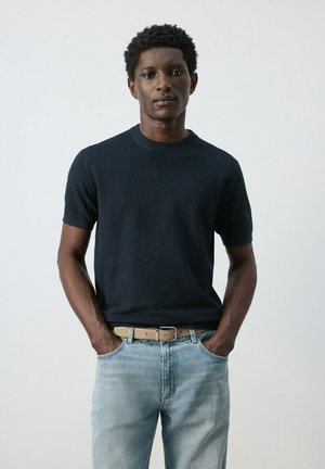 Mladi moški s temnimi kodrastimi lasmi, oblečen v črni pulover s kratkimi rokavi, svetlo modre kavbojke in bež pas, stoji z rokami v žepih.