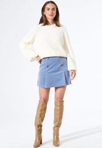 Frau in einem weißen Strickpullover, blauer Knopfleiste aus Cordstoff und kniehohen, beigen Lederstiefeln, stehend vor einem weißen Hintergrund.