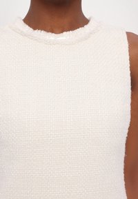 Gros plan d'un haut sans manches blanc cassé texturé avec un col rond effiloché porté par une personne à la peau foncée.