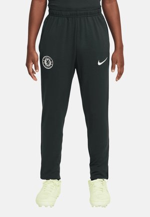 Personne portant un pantalon de sport Nike noir avec le logo du Chelsea Football Club, debout avec les bras détendus le long du corps et des crampons de couleur claire.
