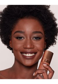 Flacone di fondotinta airbrush, con un tappo dorato rigato e un corpo liscio color beige, tenuto in mano. Il modello mostra un sorriso radioso e una pelle marrone intensa.