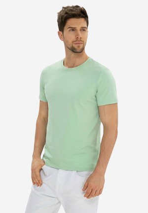 Uomo con capelli corti che indossa una maglietta verde chiaro a maniche corte e pantaloni bianchi, guarda leggermente di lato, mani rilassate.