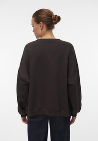 Vrouw met een bun in haar haar, gekleed in een donkerbruine oversized trui en donkere jeans, met de rug naar de camera gekeerd tegen een effen witte achtergrond.