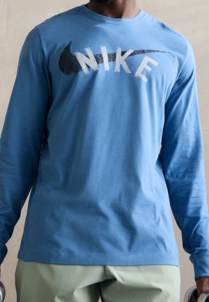 Mand iført en lyseblå langærmet skjorte med et falmet sort Nike-swoosh og hvid Nike-tekst på brystet.