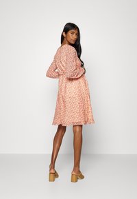 Vestido floral em laranja pastel com mangas longas, costas franzidas e comprimento até aos joelhos. Combinado com mules de salto bloco em bege.