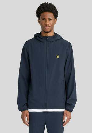 Junger Mann trägt eine marineblaue Kapuzenjacke mit Reißverschluss und kleinem gelben Vogel-Emblem auf der Brust, steht vor einem einfarbigen hellen Hintergrund.