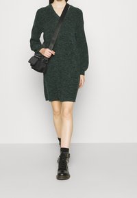 Mörkgrön stickad klänning med V-ringning och långa ballongsärmar, kombinerad med en svart crossbody-väska och svarta snörstövlar.