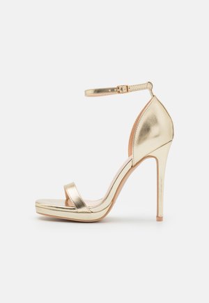 Guldmetallic højhælet sandal med ankelrem, åbent tådesign og en platformsål. Har en glat tekstur og en elegant silhuet.