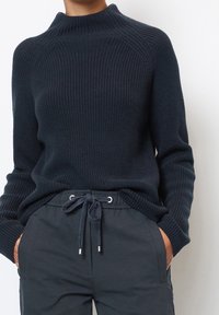 Maglione a coste di un blu navy scuro con colletto alto e maniche a raglan, abbinato a pantaloni con coulisse scuri dotati di dettagli in metallo.