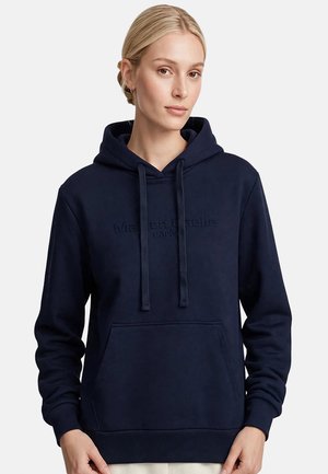 Mujer rubia con sudadera azul marino con texto en relieve, de pie frente a un fondo blanco liso.