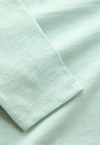 Tissu vert menthe clair avec une texture douce. Gros plan d'un ourlet de manches, mettant en avant des détails de couture et une finition lisse.