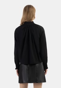 Schwarze Bluse mit langen Ärmeln, hohem Kragen, gerafftem Rückendetail und elastischen Bündchen, kombiniert mit einem engen schwarzen Lederrock.
