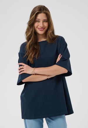Ung kvinde med langt brunt hår smiler, iført en oversized navyblå skjorte og lyseblå jeans, armene krydsede, foran en ensfarvet lys baggrund.