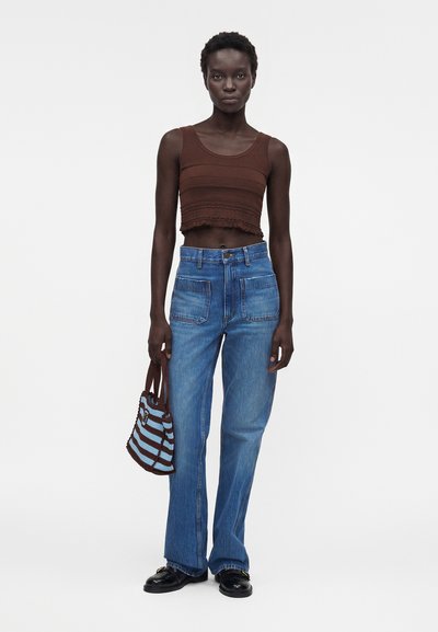 Γυναίκα με καφέ αμάνικο cropped τοπ, ψηλόμεσο μπλε τζιν, μαύρα loafers, κρατώντας καφέ και μπλε ριγωτή τσάντα.