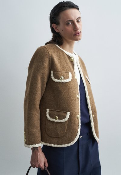 Veste en laine marron avec une finition texturée, passepoil blanc, boutons dorés et deux poches avant. Le modèle tient un sac à main marron.
