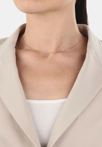 Roséguld chokerhalsband med en delikat kedja och små cirkulära detaljer, bärs på en bar nyckelben, matchad med en beige kavaj.