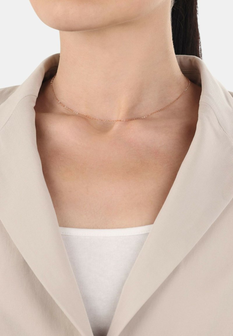 Roséguld chokerhalsband med en delikat kedja och små cirkulära detaljer, bärs på en bar nyckelben, matchad med en beige kavaj.