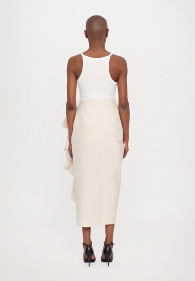 Jupe couleur crème avec un accent en volants, associée à un débardeur blanc. Des chaussures à talons hauts noires complètent la tenue. Design minimaliste.