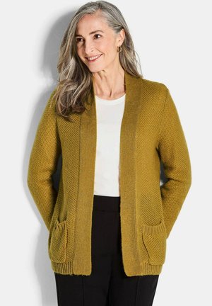 Cardigan jaune moutarde avec un motif tricoté texturé, à l'avant ouvert, et deux poches avant. Porté sur un haut blanc et un pantalon noir.
