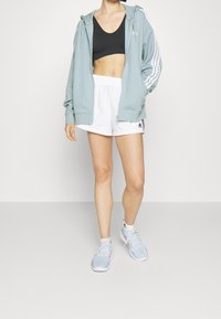 Světlá modrá mikina na zip, černá sportovní podprsenka, bílé trenýrky s černými detaily a světlé modré sportovní boty s růžovými detaily.