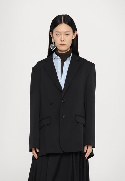 Blazer nero doppiopetto con design strutturato, dotato di due tasche frontali e bottoni neri; indossato su una camicia a righe azzurra chiara.