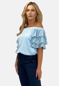 Jasnoniebieska bluzka typu off-the-shoulder z warstwowymi falbanami na rękawach, wykonana z materiału podobnego do satyny, w zestawie z dopasowanymi, ciemnoniebieskimi spodniami.