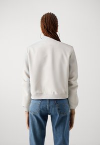 Veste bomber gris clair avec col et poignets côtelés, en tissu texturé, associée à un jean en denim bleu. Les détails incluent des poches latérales et une silhouette épurée.