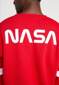 Felpa rossa con grande scritta bianca "NASA" sulla schiena. Il tessuto è un misto cotone, con scollatura rotonda e maniche lunghe.