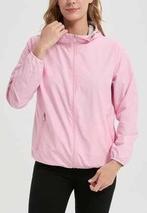 Femme ajustant le col d'une veste zippée rose clair avec poignets élastiques et poches latérales zippées, portant un pantalon noir.