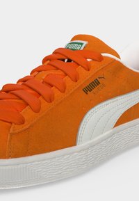 Πορτοκαλί sneaker Puma από σουέντ με λευκή ρίγα, πορτοκαλί κορδόνια και υφασμένη λευκή καουτσούκ σόλα σε ουδέτερο φόντο.
