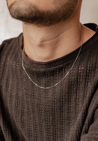 Collana sottile in argento appoggiata su una maglietta nera a maglia testurizzata, che mostra un design semplice e curvo con una lucentezza sottile.
