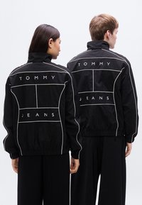 Μαύρο αθλητικό μπουφάν με λευκά γραφικά περιγράμματα και τα κείμενα "TOMMY" και "JEANS" στην πλάτη, με ψηλό γιακά και ελαστικές μανσέτες.