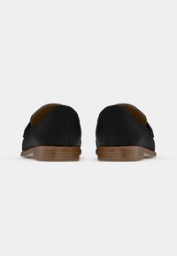 Pantofi loafer din piele neagră, cu vârf rotunjit, accente cusute și tălpi plate din lemn, văzuți din spate. Textură netedă, design minimalist.