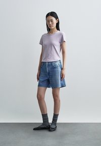 Fialové tričko s krátkym rukávom, svetlo modré rifľové šortky, sivé rebrované ponožky a čierne baleríny, stojace na sivom podlaží pred jednofarebným pozadím.