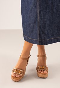 Sandalias de cuña marrones con una plataforma tejida texturizada, correa de tobillo ajustable y detalle decorativo de hebilla. Pies visibles, combinadas con una falda de mezclilla.