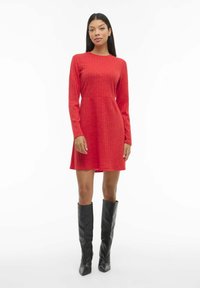 Robe rouge à manches longues avec une texture côtelée, corsage ajusté et jupe évasée. Associée à des bottes noires montant jusqu'au genou et un design minimaliste.