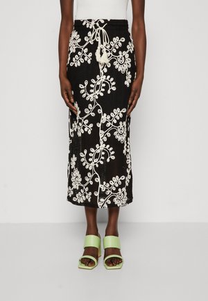 Black midi skirt with white floral embroidery, textured fabric, cinched waist with drawstring, paired with green block-heeled sandals.