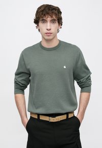 Πράσινο πλεκτό πουλόβερ με μακριά μανίκια και στρογγυλή λαιμόκοψη, με μικρό λογότυπο στο στήθος, σε συνδυασμό με ζώνη καμηλοπάρδαλης και μαύρο παντελόνι.