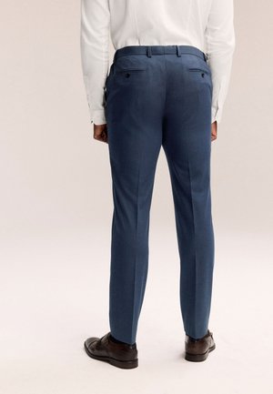 Bărbat purtând pantaloni eleganți albaștri slim-fit, cămașă albă cu mânecă lungă și pantofi eleganți din piele maro închis, văzut din spate.