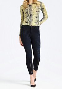 Długosleeve'owa bluzka w wzór węża w żółtym i czarnym kolorze, zestawiona z czarnymi jeansami skinny z przetarciami i zamkami błyskawicznymi na końcach nogawek.
