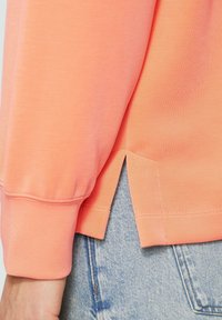 Perzikkleurig sweatshirt met lange mouwen, voorzien van een zijsplit en geribbelde cuffs, gecombineerd met lichtblauwe denim jeans.
