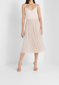 Ljusrosa ärmlös topp i kombination med en svängd, knälång tullekjol med prickar. Modell som håller en silverfärgad kuvertväska och bär hudfärgade klackar.