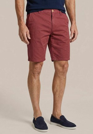 Man wearing maroon shorts, navy blue shirt, and navy blue slip-on shoes against a plain beige background.
