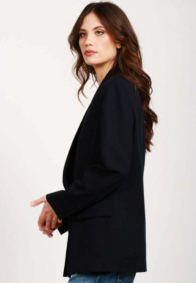 Blazer nero con un design sartoriale, rever a punta e maniche lunghe. Presenta una texture liscia e una vestibilità rilassata, abbinato a jeans blu.