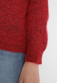 Czerwony sweter o fakturowanej powierzchni, wykonany z miękkiego materiału dzianinowego, z prążkowanymi mankietami i prostym dołem, zestawiony z jasnymi dżinsami.