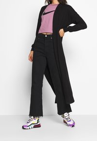 Svart lång kofta över en rosa kort t-shirt med en svart logotyp, parat med högmidjade svarta utsvängda jeans och flerfärgade sneakers.